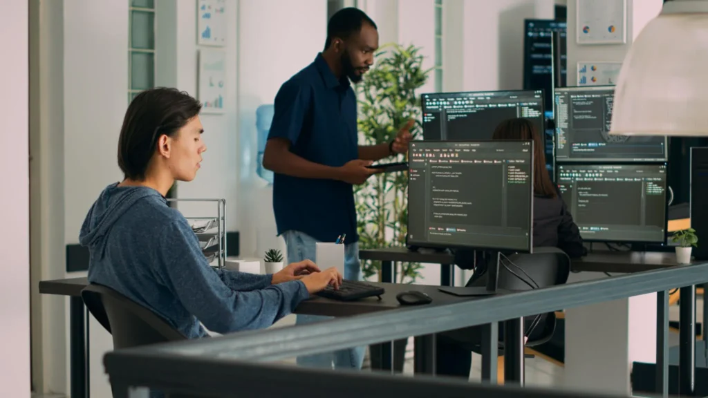 Two individuals working at desks with multiple computer monitors in a modern office setting.